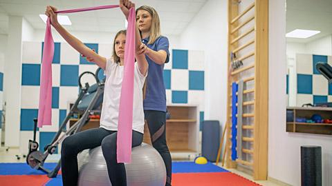 Steadied by a therapist, a atient sits on a silver ball while stretching a pink elastic band above her head. 